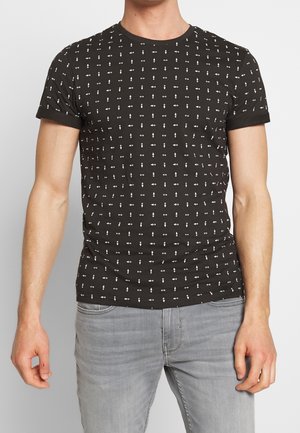 Mies, jolla on yllään musta, istuva T-paita, jossa on pieniä valkoisia geometrisia kuvioita, yhdistetty vaaleanharmaisiin farkkuihin, seisoo yksinkertaista taustaa vasten.