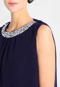 Robe sans manches bleu marine avec un col froncé, ornée d'un col en cristal. Tissu lisse avec une silhouette fluide.