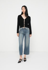 Cardigan preto cropped com um decote V profundo e detalhes em botão dourado, combinado com umas calças de ganga de perna larga azul claro e mules pretos de bico fino e salto.