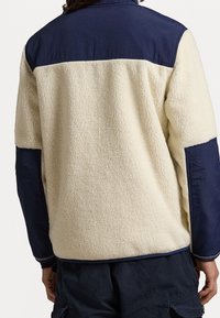 Fleece jack met een crèmekleurig lichaam en een marineblauwe bovenkant; voorzien van een hoge kraag en contrasterende mouwen, met een zachte textuur en nette naden.