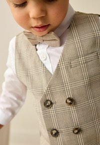 Gilet à carreaux beige avec boutons croisés, associé à une chemise blanche et un nœud papillon beige assorti. Détail de texture du tissu visible.