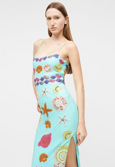 Ung kvinde iført en lyseblå kjole med farverige pailletmønstre af havdyr og muslinger, med tynde stropper og et slids til låret.