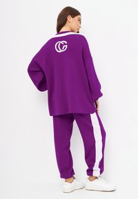 Pull en tricot oversized violet avec un logo blanc au dos, associé à un pantalon violet ajusté avec une bande blanche sur le côté.