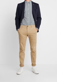 Blazer azul marino sobre una camisa de cuadros en blanco y negro, combinado con chinos beige y zapatillas blancas. Los pantalones están remangados en los puños.