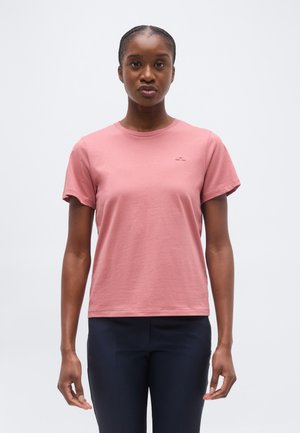 Paelustega juustega naine, kes kannab lihtsat lühikeste varrukatega roosat t-särki ja tumedaid pükse, seisab helehallil taustal.