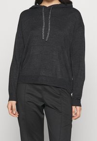 Černá mikina s kapucí a strukturovaným povrchem, s přední kapsou, stříbrnými tkaničkami a žebrovanými manžetami, nošena s odpovídajícími kalhotami.