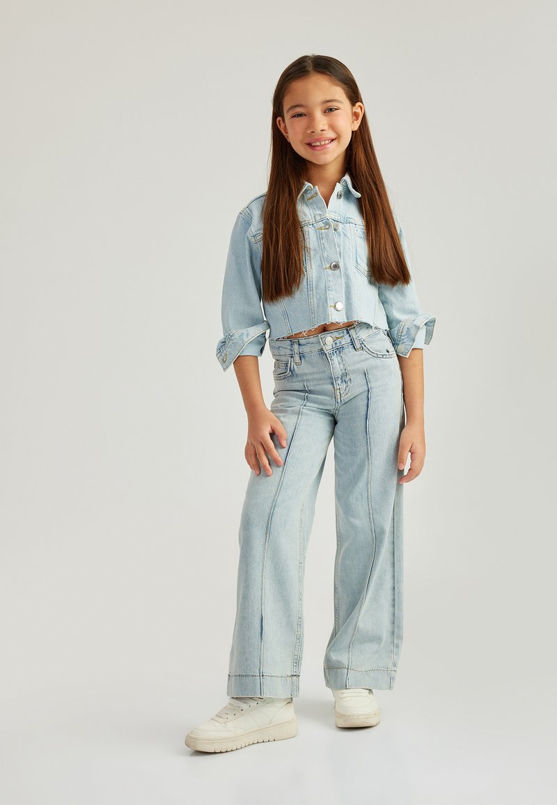 Giacca di jeans azzurro chiaro cropped con maniche arrotolate, abbinata a jeans a gamba larga alta vita e sneakers bianche. Design semplice con dettagli cuciti.