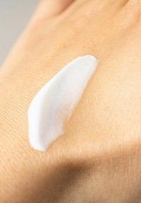 Weiße Creme auf einem hellen Hautton aufgetragen. Die Creme ist glatt und hat ein glänzendes Aussehen, welches die Hautstruktur hervorhebt.