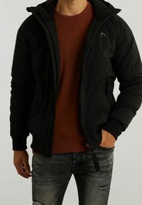Sort isoleret jakke med lynlås foran, hætte og struktureret stof. Bæres over en rød sweater og grå slidte jeans.