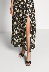 Czarna maxi sukienka w kwiaty z żółtymi i białymi elementami, z bocznym rozcięciem. Noszona z czarnymi sandałami na obcasie z odkrytymi palcami.