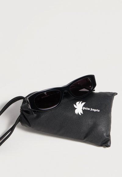 Lunettes de soleil rectangulaires noires posées sur une pochette noire à cordon avec un logo de palmier blanc et le texte « Palm Angels ».