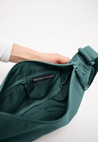 Hand hält eine offene türkisfarbene Stofftasche, die eine Reißverschlusstasche innen mit einem Etikett zeigt, auf dem "FROM TRASH TO TREASURE." steht