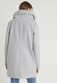 Kobieta ubrana w jasnoszary wełniany płaszcz z dużym złożonym kołnierzem, pokazana z tyłu na jednolitym tle.