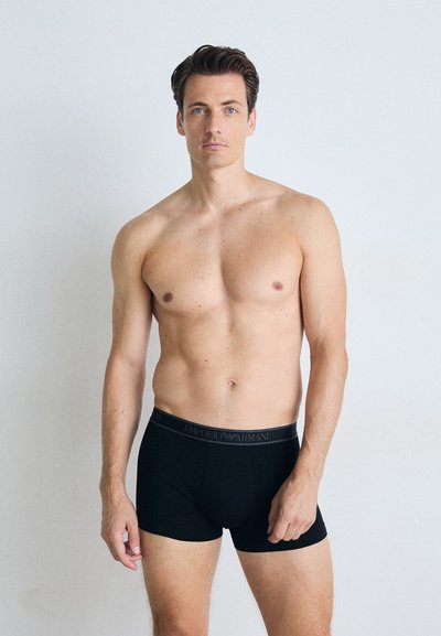 Crni pamučni bokserici s elastičnom gumom koja sadrži "Emporio Armani" logo. Model stoji u neutralnom okruženju.
