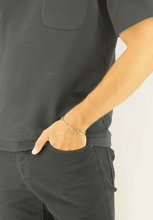 Schwarzes Kurzarmhemd mit Brusttasche, kombiniert mit schwarzen Hosen. Ein goldfarbenes Gliederarmband ist am Handgelenk zu sehen und sorgt für einen kontrastierenden Akzent.