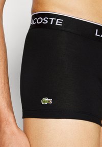 Schwarze Baumwollboxershorts mit einem Lacoste-Logo-Bund und einem kleinen grünen Krokodil, das auf der unteren linken Seite gestickt ist.