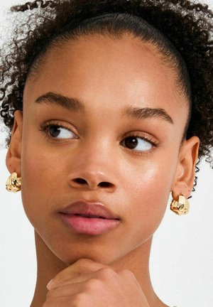 Kullavärvi metallist kõrvarõngad abstraktse disainiga, millel on sile ja peegeldav pind. Modelli juuksed on lokkis ja meik loomulik.