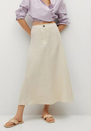 Vrouw die een beige midi-rok met hoge taille draagt, een lavendelkleurige cropped blouse met opgerolde mouwen en beige geplooide platte sandalen.