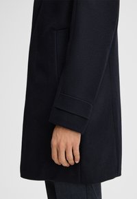 Donkerblauwe jas met een gladde textuur, voorzien van een zijzak en een omgeslagen manchetdetail. Het ontwerp heeft een rechte snit voor een getailleerde look.