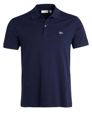 Homme marchant portant un polo bleu marine, un short beige, des chaussettes blanches et des baskets noires sur un fond clair uni.
