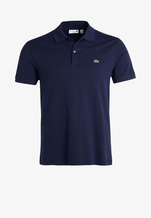 Homme marchant portant un polo bleu marine, un short beige, des chaussettes blanches et des baskets noires sur un fond clair uni.