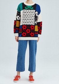 Pull coloré en maille avec divers motifs, comprenant des fleurs et des formes géométriques, associé à un jean large en denim bleu et des baskets rouges.