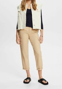 Kvinna klädd i beige cargobyxor med uppvikta benslutar, svart långärmad topp, off-white ärmlös knäppt väst och svarta slidessandaler.