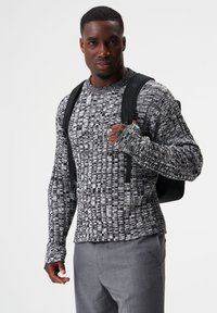 Strickpullover in Schwarz-Weiß mit Struktur, Rundhalsausschnitt, langen Ärmeln und lockerem Schnitt, kombiniert mit grauen Hosen und einem schwarzen Rucksack.