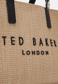 Gros plan sur un sac à main tissé beige avec des anses en cuir noir et le logo brodé "TED BAKER LONDON" en noir.