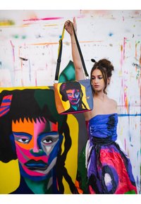 Bunte Tragetasche mit einem auffälligen Grafikdesign eines Gesichts. Hergestellt aus glattem Material, mit einem schwarzen Trageriemen und gelben Akzenten.