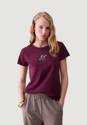 Mujer joven con cabello rubio que lleva una camiseta morada con iniciales y pantalones beige, de pie con las manos en los bolsillos frente a un fondo liso.