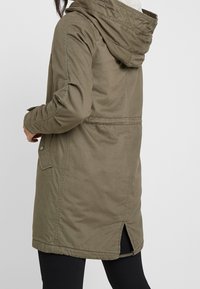 Olivgrüne Parka mit Kapuze, ausgestattet mit einem gesteppten Design, seitlichen Taschen und einem Rückenschlitz für bessere Bewegungsfreiheit. Hergestellt aus weichem, langlebigem Material.