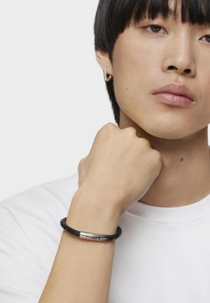 Pulsera de cuero tejida negra con un cierre cilíndrico de plata. La mano descansa cerca del cuello, llevando una camiseta blanca y un pendiente de aro plateado.