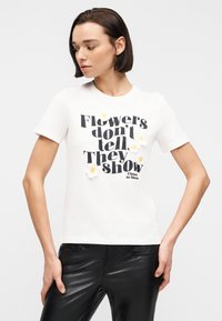 Γυναίκα με κοντά σκούρα μαλλιά που φοράει λευκό μπλουζάκι με μαύρο κείμενο «Τα λουλούδια δεν λένε, δείχνουν» και μικρές λευκές μαργαρίτες κεντημένες.