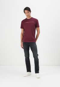 Burgunderfarbenes Baumwoll-T-Shirt mit kurzen Ärmeln und Rundhalsausschnitt, mit weißem "TOMMY JEANS"-Logo auf der Brust, kombiniert mit dunklen Jeans und weißen Sneakern.