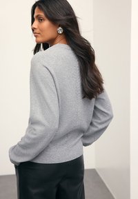 Gray knit sweater with a textured pattern and ribbed hem. Model shown from the back, paired with black pants. Circular earrings visible.