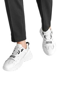 Baskets blanches avec des superpositions en maille et en cuir, portant des accents noirs et un logo "VERSACE JEANS COUTURE" sur les côtés. Semelles en caoutchouc épaisses.