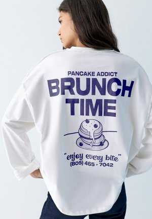 Mladá žena v bielej košeli s dlhým rukávom s tmavomodrým nápisom "Pancake addict BRUNCH TIME" a ilustráciou palacinky na chrbte.