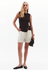Top senza maniche nero con dettagli in bottone dorato, abbinato a pantaloni corti color crema. La modella tiene una piccola borsa nera e indossa delle ballerine nere.