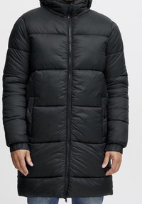 Zwarte pufferjas met een capuchon, voorzien van een ritssluiting aan de voorkant, elastische manchetten en een horizontaal gestikt patroon. Lengte tot halverwege de bovenbenen met zijzakken.