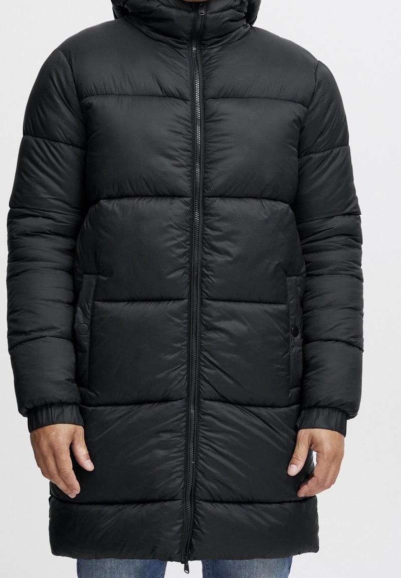 Zwarte pufferjas met een capuchon, voorzien van een ritssluiting aan de voorkant, elastische manchetten en een horizontaal gestikt patroon. Lengte tot halverwege de bovenbenen met zijzakken.