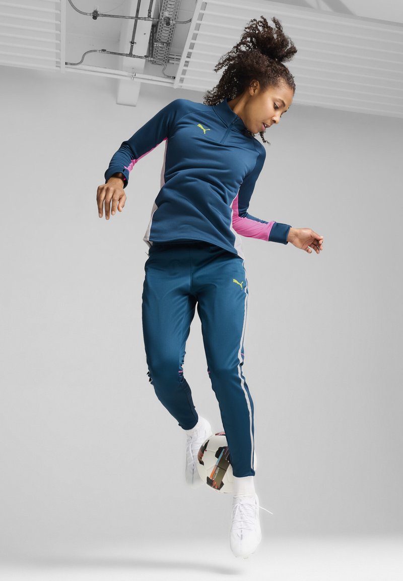 Tuta sportivo teal con accenti rosa e righe bianche. Il modello, in volo, indossa scarpe bianche e interagisce con un pallone da calcio.