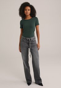 Vrouw met krullend haar, gekleed in een donkergroene shirt met korte mouwen, hooggeknoopte grijze wijde broek met een strik riem, en zwarte puntige schoenen.
