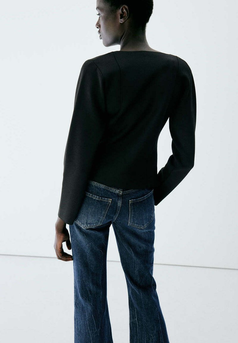 Personne portant un haut noir à manches longues et un jean bleu en denim, debout de profil avec une main posée sur la hanche.