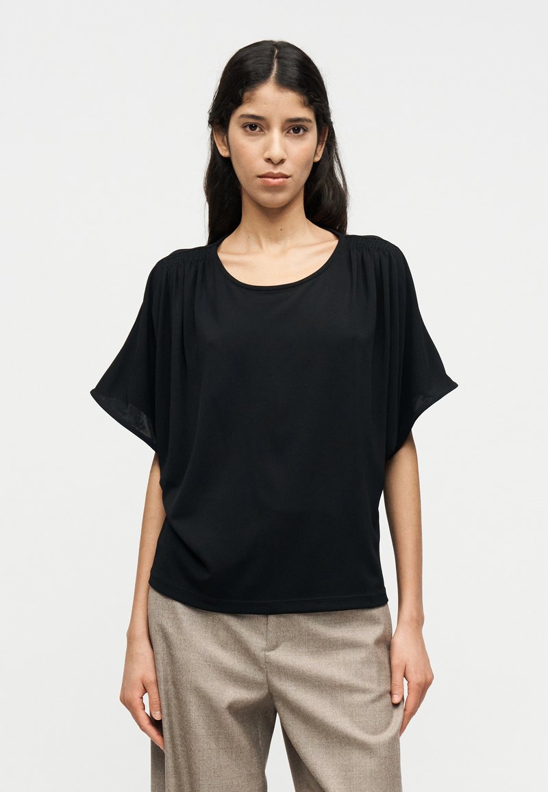 Μαύρο μπλουζάκι με κοντά μανίκια από ελαφρύ ύφασμα με στρογγυλή λαιμόκοψη και πιέτες στους ώμους. Συνδυάζεται με μπεζ παντελόνια.