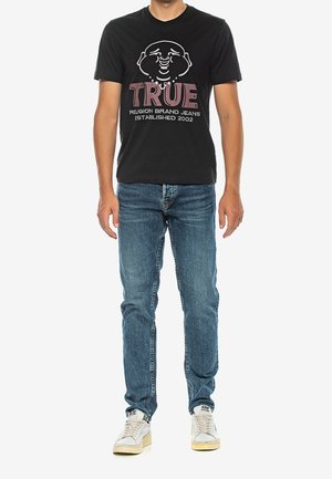 Hombre con camiseta negra con dibujo de cara de caricatura y texto "TRUE Religion Brand Jeans Established 2002", jeans azules y zapatillas blancas, de pie contra fondo blanco.