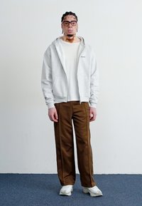 Grå zip-up hoodie med lommer, hvid t-shirt, brune brede bukser og hvide sneakers. Ensfarvet baggrund med neutrale farver.