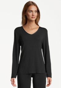 Young woman with light brown hair wearing a black long-sleeve V-neck top and matching black pants, facing forward against a white background.