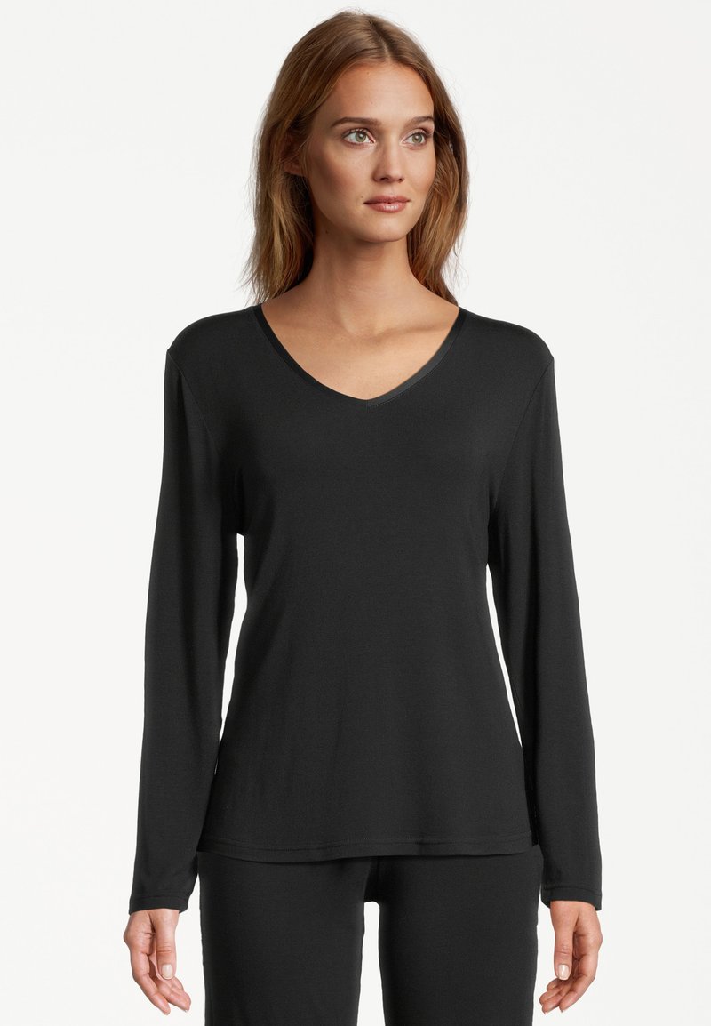 Young woman with light brown hair wearing a black long-sleeve V-neck top and matching black pants, facing forward against a white background.