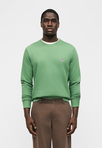 Νέος άνδρας φορά πράσινο ανοιχτό φούτερ με μικρό λογότυπο ζέβρας στο στήθος και καφέ παντελόνια, στέκεται μπροστά σε απλό φόντο.